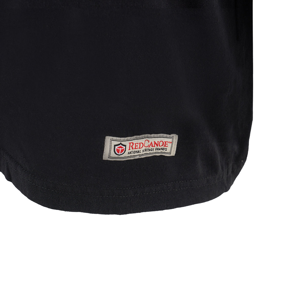 Close-up of the hem of a black Red Canoe Boeing Defense T-Shirt by The Boeing Store, featuring a small rectangular patch label with a red canoe logo—a nod to WW2 era heritage.