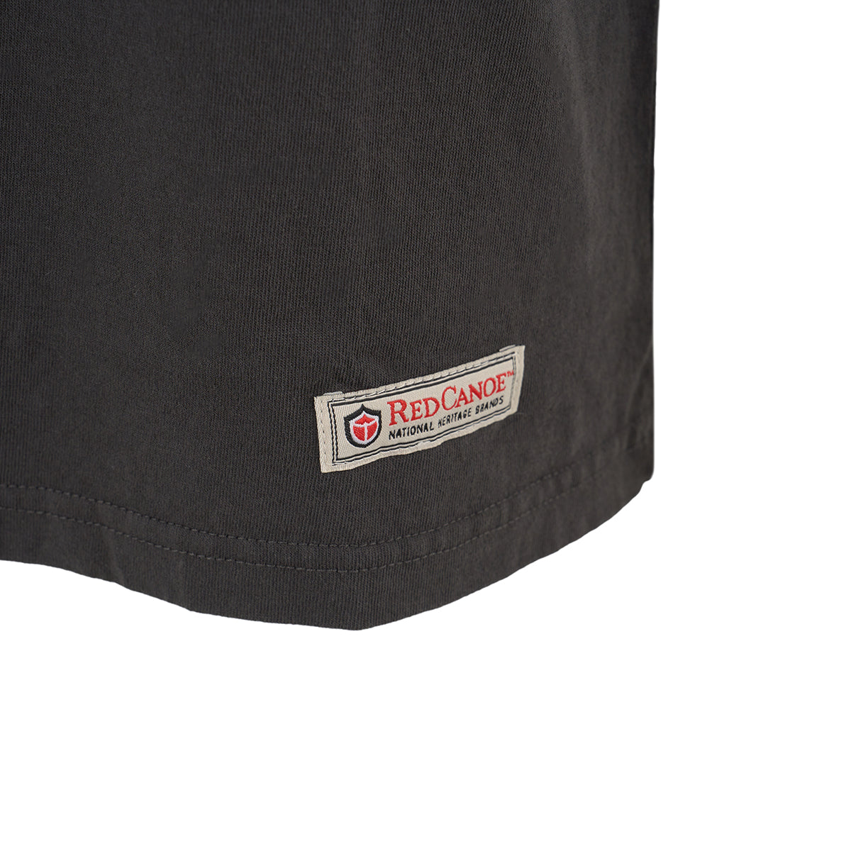 Close-up of the lower hem of a dark Red Canoe Boeing Tuskegee Airmen T-Shirt from The Boeing Store, featuring a rectangular patch with Red Canoe National Heritage Brands and a red shield logo, inspired by the Tuskegee Airmen.