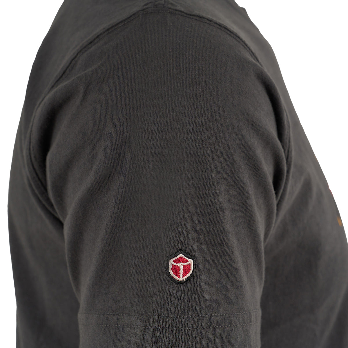 Close-up of a person’s upper arm wearing The Boeing Store’s black Red Canoe Boeing Tuskegee Airmen T-Shirt, featuring a small red and white shield-shaped logo on the sleeve. Plain white background.