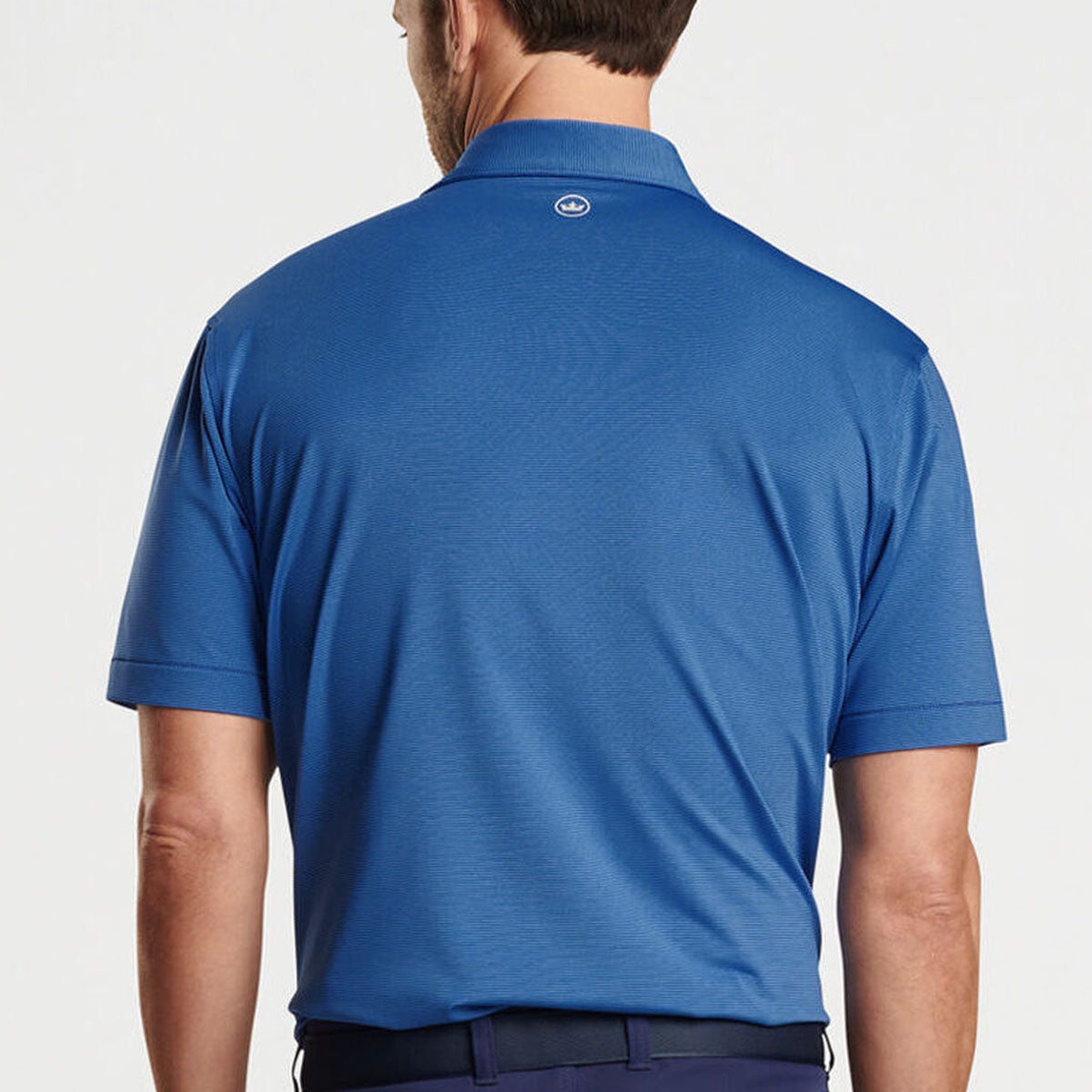 A man is seen from behind wearing the Peter Millar Boeing Men’s Jubilee Performance Polo in blue with 4-way stretch, featuring a small circular Boeing logo below the collar, paired with dark pants.