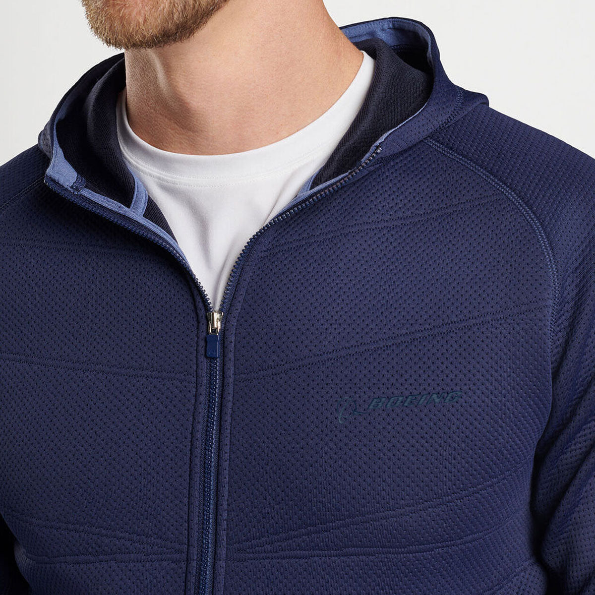 A man wears a white T-shirt and the Peter Millar Boeing Men’s Orion Performance Quilted Hoodie in navy blue, featuring subtle chest branding, a textured perforated design, and four-way stretch. Only his lower face and upper torso are visible.
