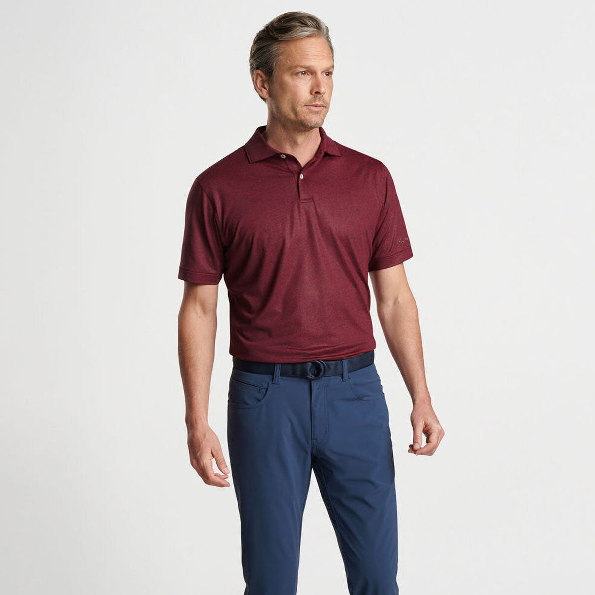 A man stands against a white background, wearing the Boeing Peter Millar Men’s Switching Gears Performance Polo in maroon with short sleeves and blue pants, looking slightly to the side with a neutral expression.