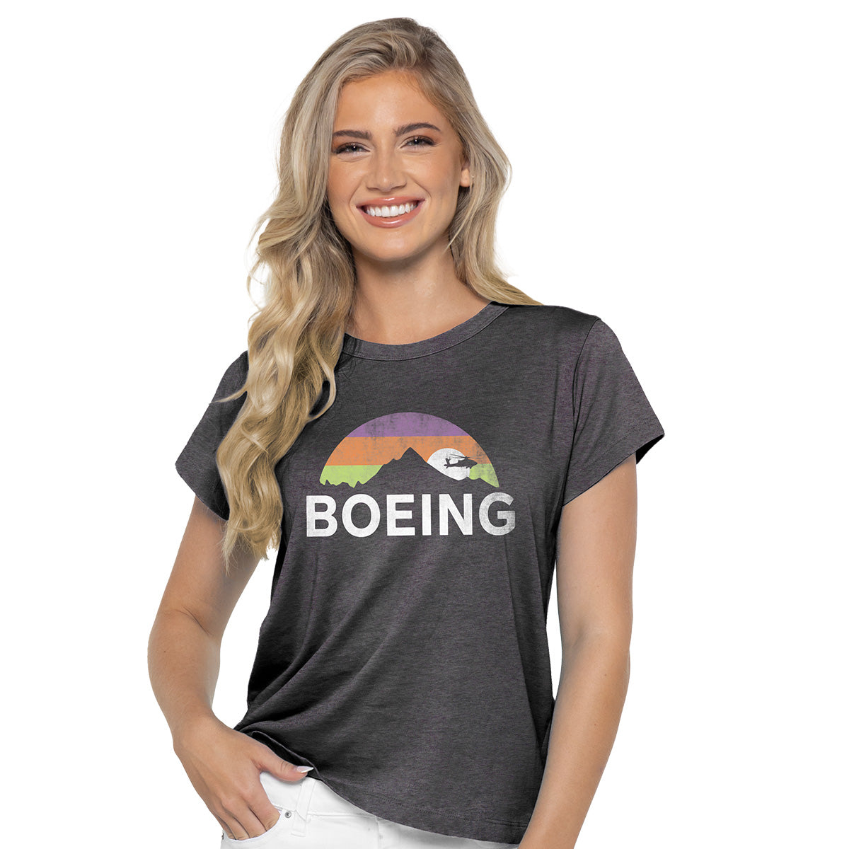 A smiling woman with long blonde hair wears a Boeing Horizon Peak Womens T-Shirt in dark gray, featuring a rainbow landscape and the Boeing logo with a stylized plane. She exudes effortless style, casually placing one hand in her pocket.