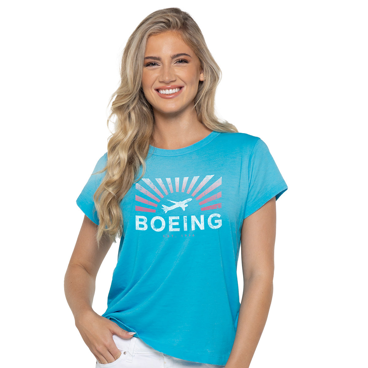A woman with long blonde hair wears the Boeing Sunbeam Womens T-Shirt by Boeing, featuring an airplane, sunbeams, and BOEING text. Ideal for aviation enthusiasts, she poses against a plain white background with one hand in her pocket.