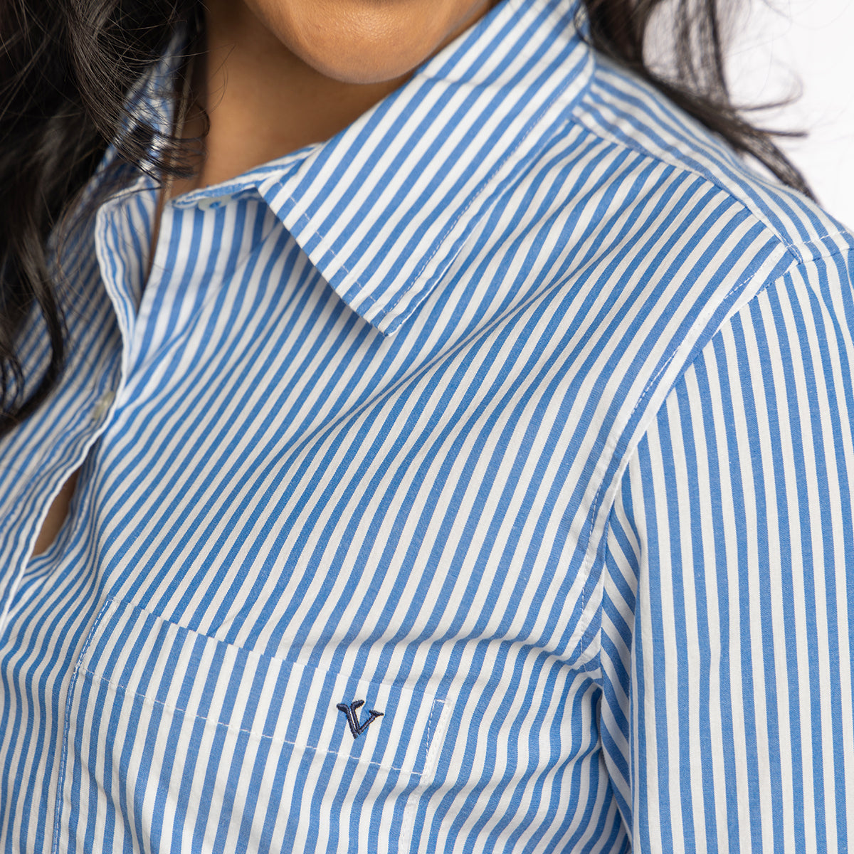 Close-up of a person wearing the Boeing Vineyard Vines Women’s Bayview Button-Down Shirt, featuring blue and white vertical stripes, premium stretch poplin fabric, a pointed collar, and an embroidered chest pocket logo.