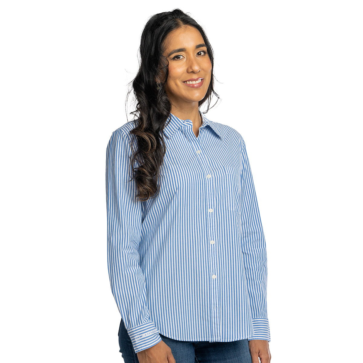 A woman with long dark wavy hair wears a Boeing Vineyard Vines Women’s Bayview Button-Down Shirt in blue and white stripes with jeans, smiling and standing against a white background.