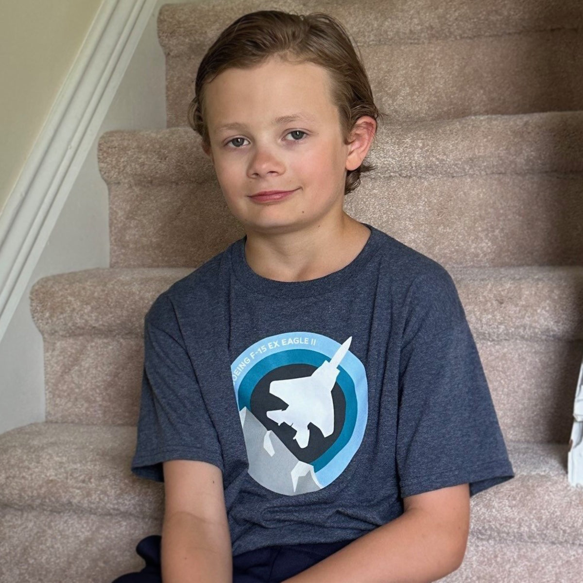 A young boy with combed-back brown hair sits on carpeted stairs, wearing a gray Boeing F-15EX Eagle Skyward Kids T-Shirt featuring a plane graphic. He smiles slightly at the camera. The neutral-colored setting enhances his charming moment in the aviation-inspired tee by Boeing.