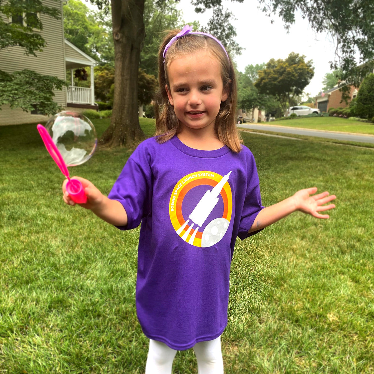 A young girl in a Boeing Space Launch System Skyward Kids T-Shirt, showing off a rocket design, waves a bubble wand on a grassy lawn. Surrounded by trees and houses, her imagination soars like the Boeing Space Launch System itself.