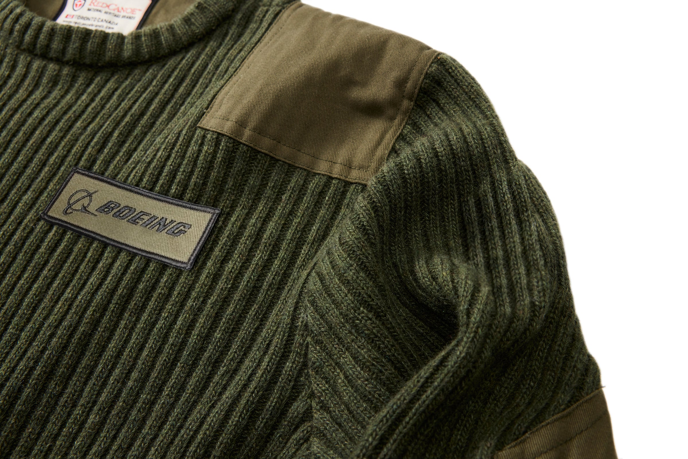 Close-up of the Red Canoe Boeing Ribbed Crewneck Sweater from The Boeing Store in olive green, featuring fabric patches on the shoulder and arm, plus a rectangular Boeing logo patch on the chest. Perfect for layering; shown against a white background.