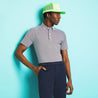 Product image on a male model leaning on a blue wall with the shirt tucked in.  Male is also wearing navy chino pants with a lime green trucker hat.
