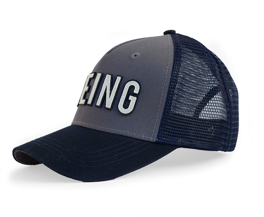 The Boeing Store’s Red Canoe Boeing Trucker Hat features navy and gray cotton twill, mesh back panels, and bold white BEING lettering on the front, inspired by classic Boeing style.