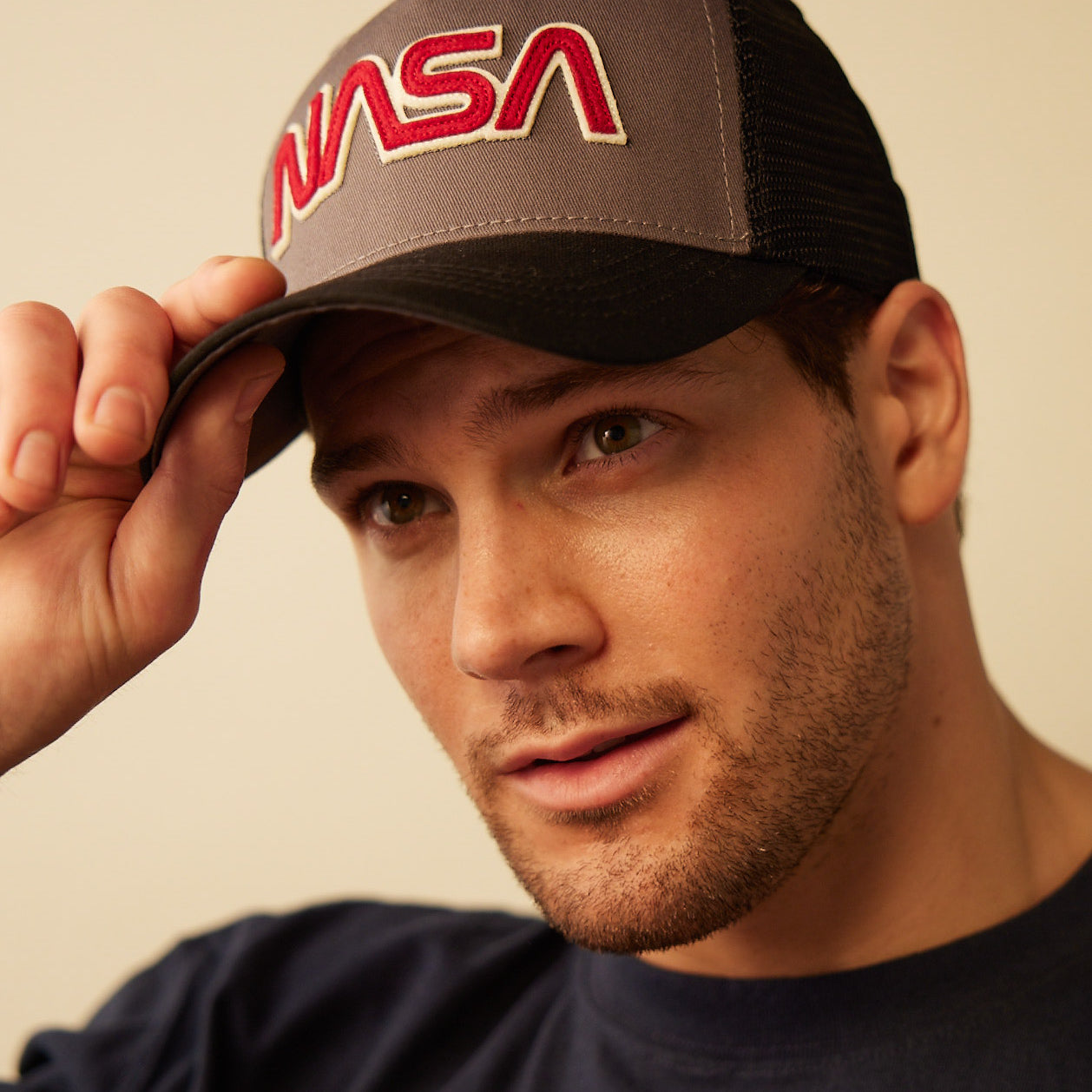 A man with light skin and a short beard wears a Red Canoe NASA Trucker Hat by The Boeing Store, black and gray with a snapback closure, looking upward while holding the brim with his right hand.