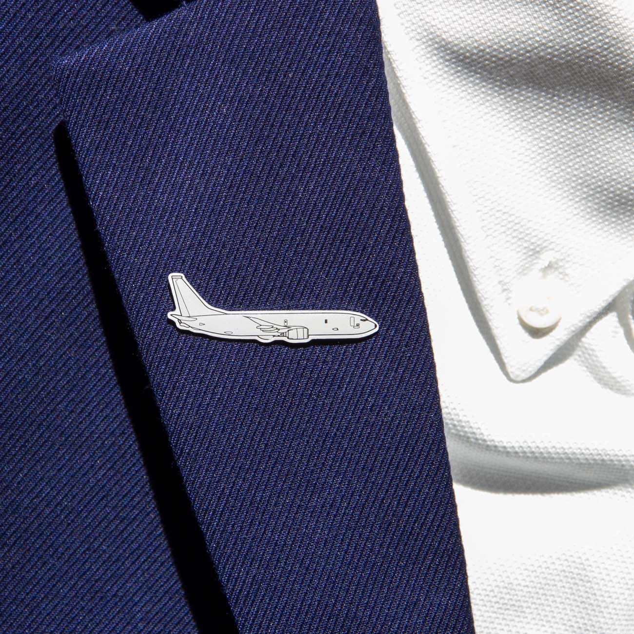 A person wears a dark blue suit jacket featuring the Boeing Illustrated P-8 Lapel Pin by The Boeing Store, shaped like an airplane and resembling the Boeing logo, on the lapel. A white textured shirt completes the look.