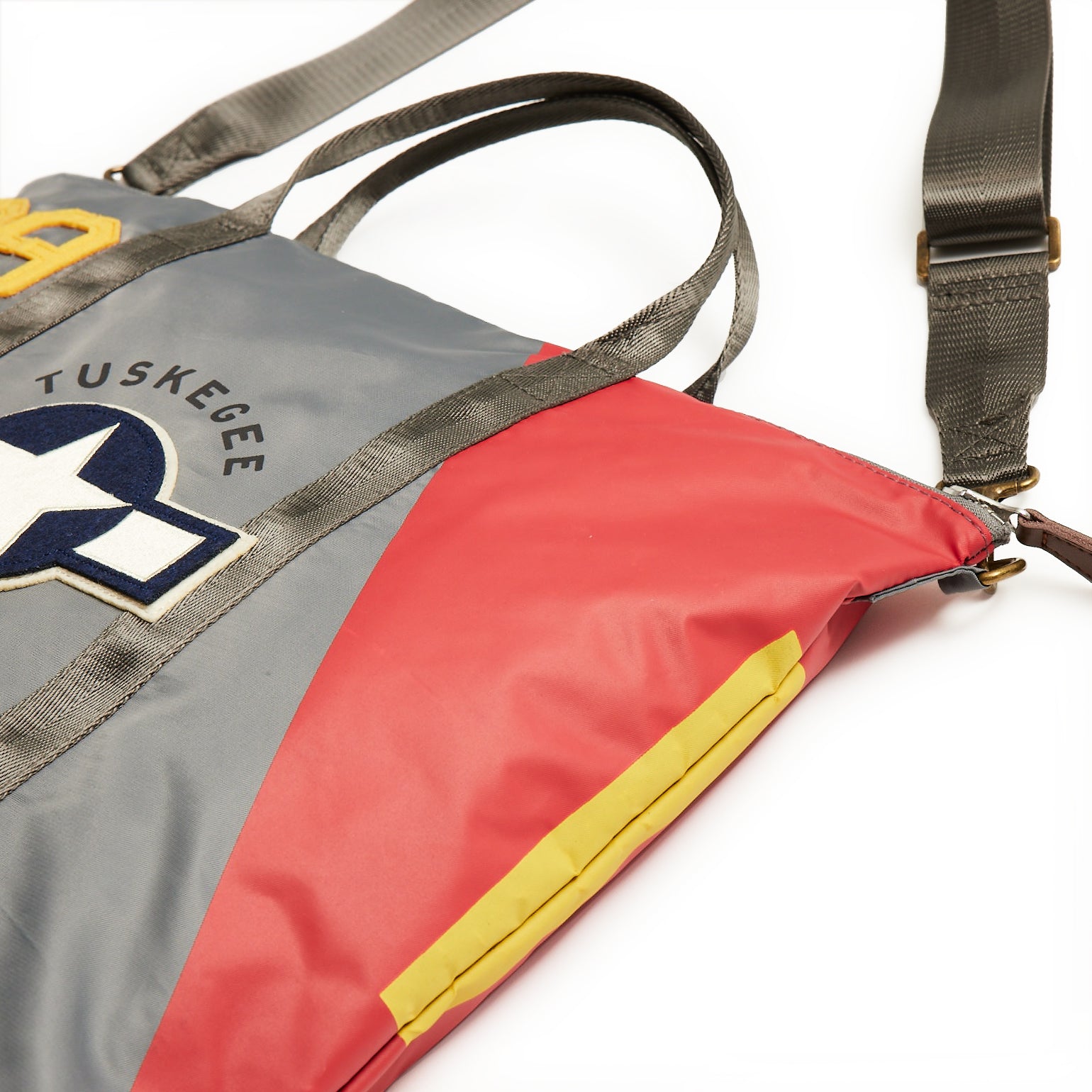 The Boeing Stores Red Canoe Tuskegee Helmet Bag is a stylish tote with the Tuskegee logo, aircraft insignia, YKK zippers, and a gray-red color scheme with yellow accents. It features sturdy handles and an adjustable shoulder strap for versatility.
