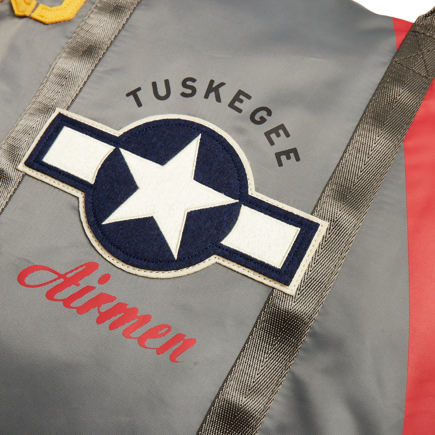 Close-up of the Red Canoe Tuskegee Helmet Bag by The Boeing Store, featuring gray fabric with a TUSKEGEE AIRMEN patch and blue star emblem. Red and yellow accents decorate the shoulder area, along with an adjustable shoulder strap for versatility.