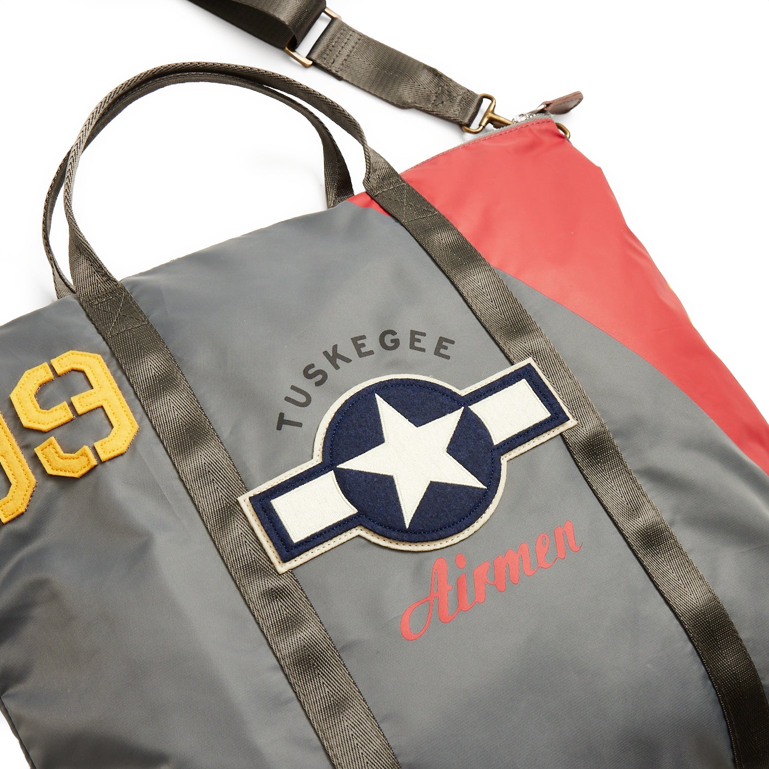 The Boeing Stores Red Canoe Tuskegee Helmet Bag is a gray and red bag with black straps, featuring embroidered patches: Tuskegee Airmen with a star insignia and 99 in yellow. It includes an adjustable shoulder strap for convenience.