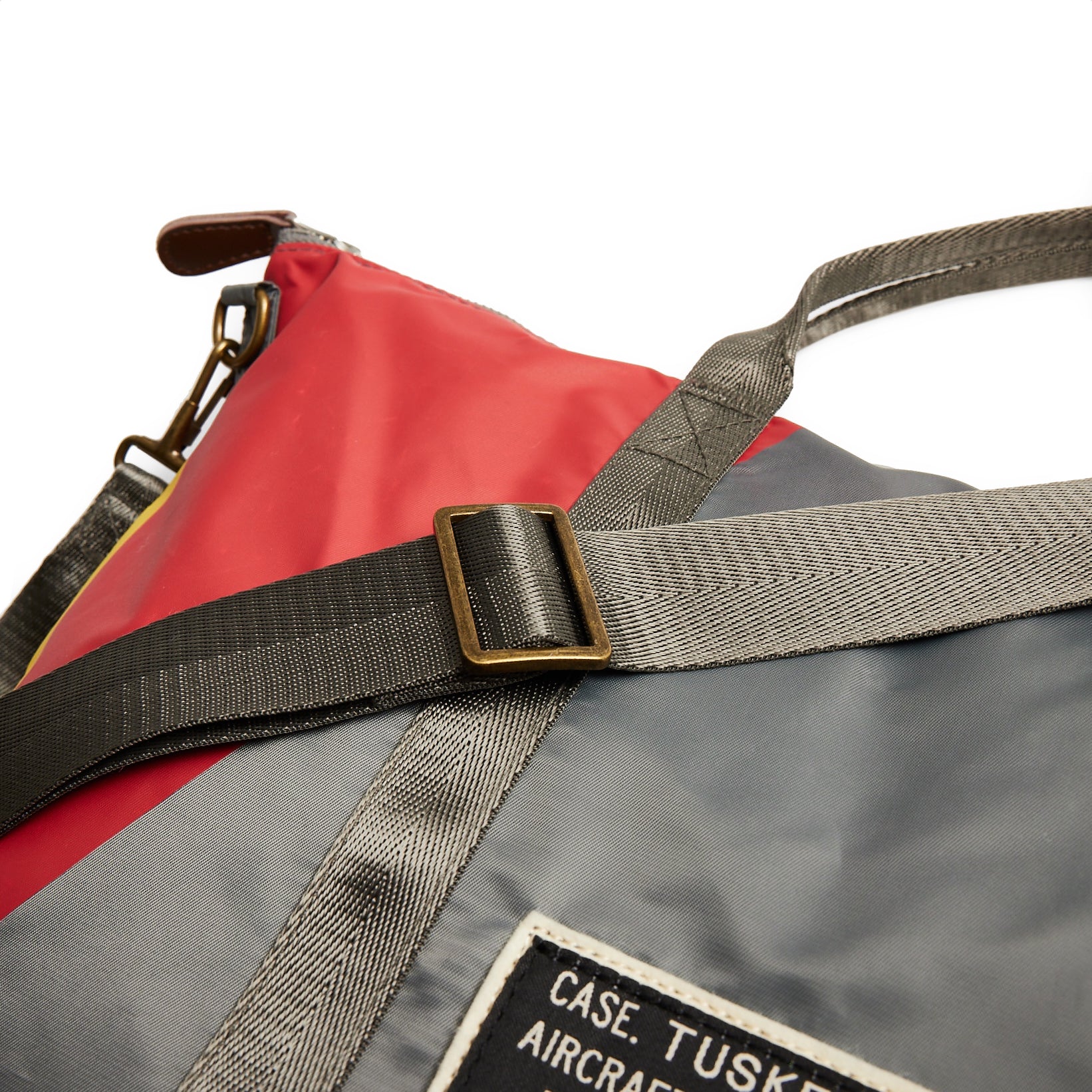 The Boeing Stores Red Canoe Tuskegee Helmet Bag features multicolored panels, including red and gray, a black adjustable shoulder strap with a metal buckle, YKK zippers, and a leather tag with text.