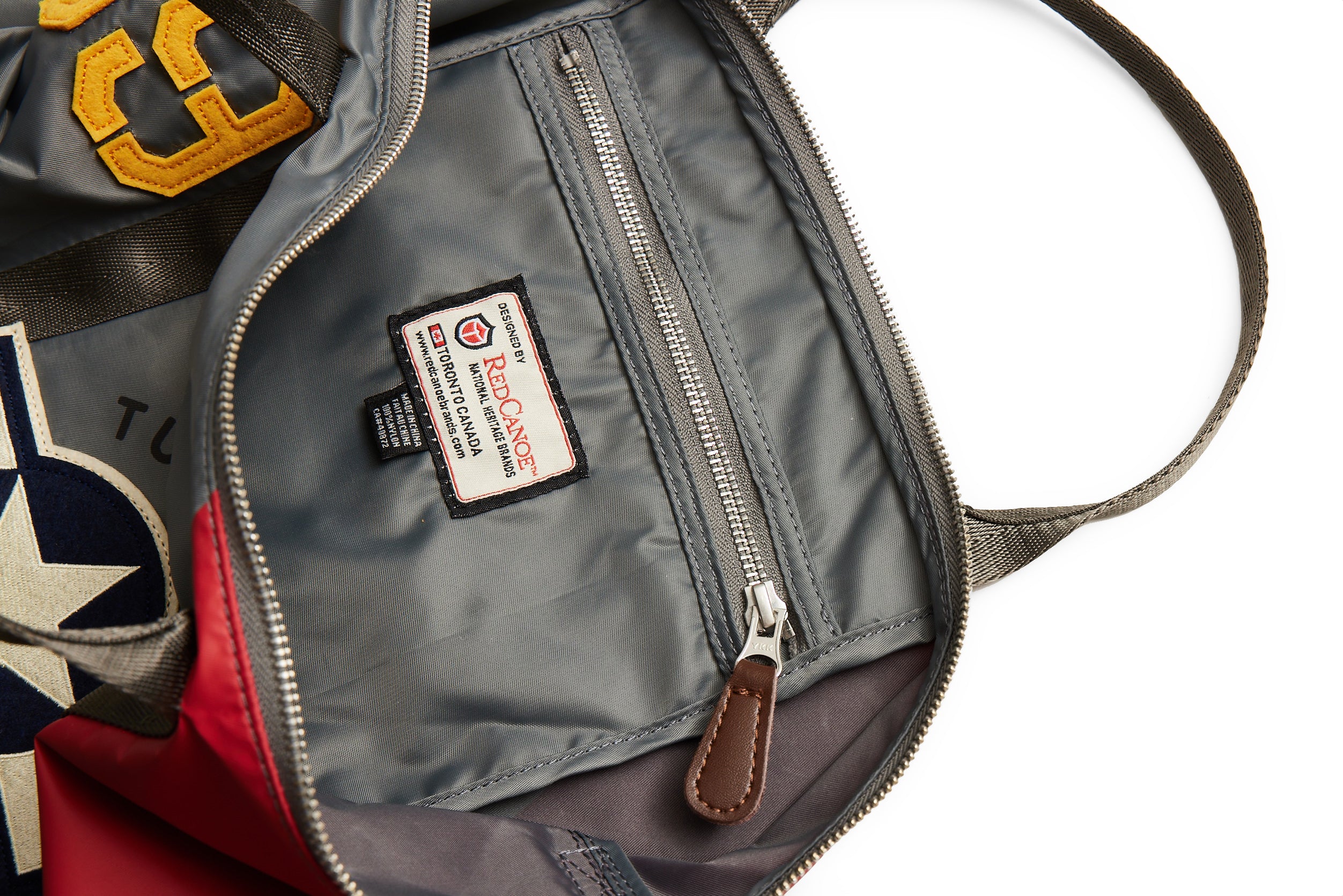 The Red Canoe Tuskegee Helmet Bag by The Boeing Store is open to reveal its interior with a zippered pocket and YKK zippers, featuring a Red Canoe label. The exterior has patches like a yellow number 8 and roundel, plus an adjustable strap for comfort, highlighting the bags design and details.