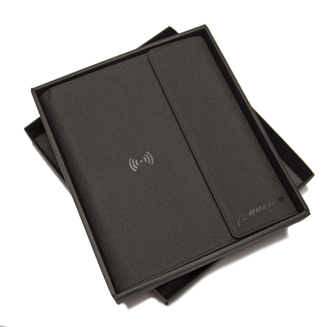A Boeing Large Padfolio With Wireless Power Bank from The Boeing Store rests in an open box. Its sleek, black faux leather cover features a wireless charging symbol and an embossed Boeing logo, combining sophistication with modern functionality.