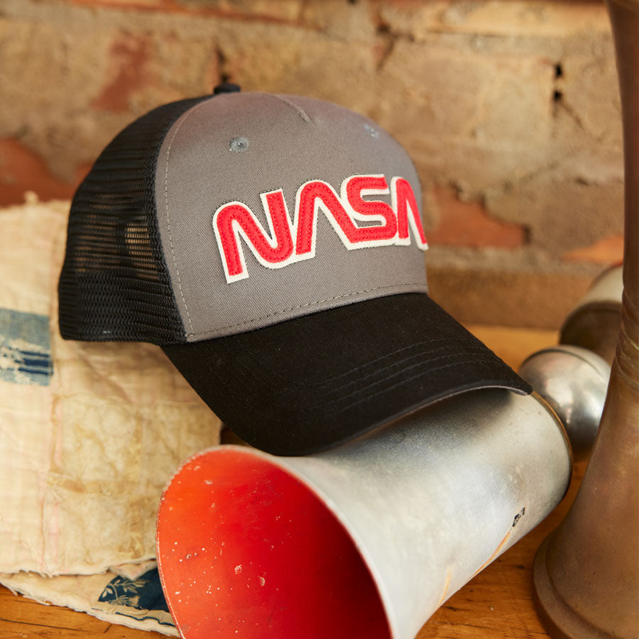 The Boeing Store’s Red Canoe NASA Trucker Hat, in gray and black with red and white embroidery, sits on a wooden surface beside a weathered metal funnel and rustic brick wall.
