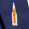 The Boeing Illustrated SLS Lapel Pin features a rocket with an orange body and white boosters on a textured dark blue fabric, reminiscent of space-inspired artwork, enhanced with silver tone sophistication.
