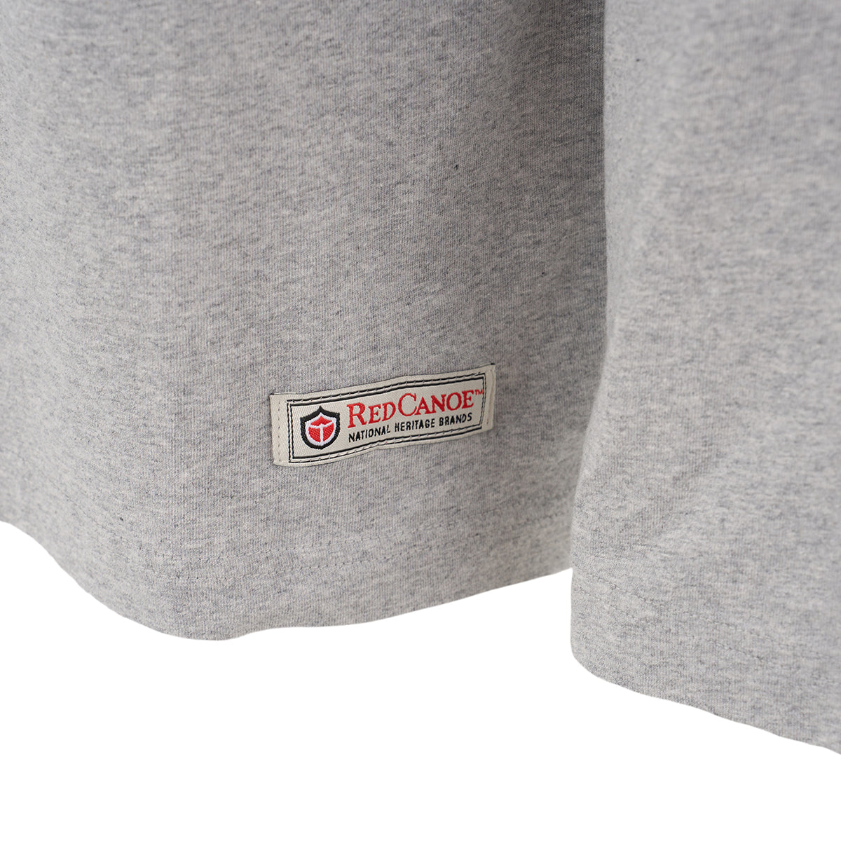 Close-up of the bottom hem of a grey Red Canoe Boeing Airplane Company Logo B-17 Long-Sleeve T-Shirt from The Boeing Store, featuring a rectangular Red Canoe patch reading “Red Canoe National Heritage Brands” stitched near the edge.