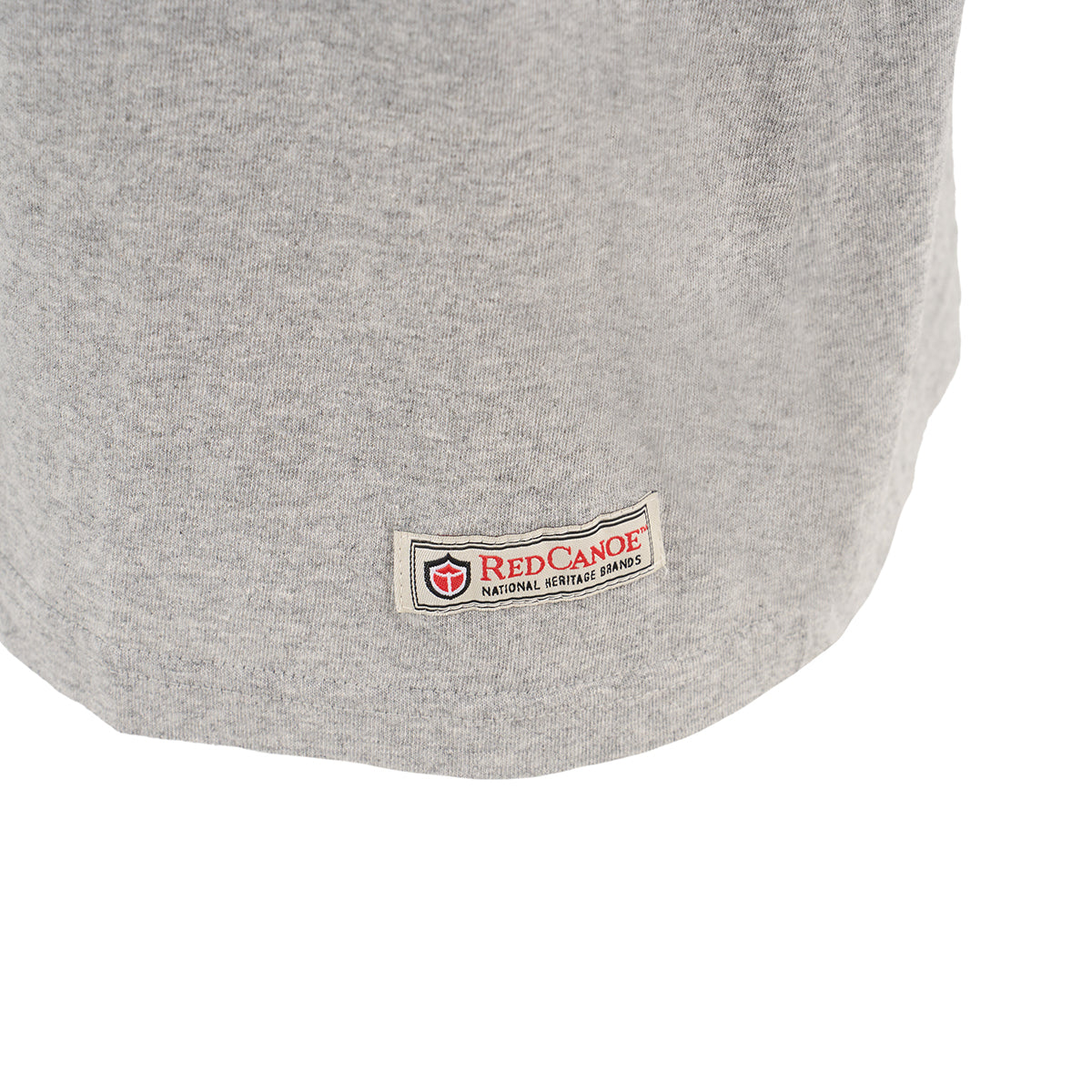 A close-up of the bottom hem of a light gray Red Canoe Boeing Airplane Company Logo B-17 T-Shirt by The Boeing Store, featuring a rectangular Red Canoe patch with red and black text and a small red shield logo.