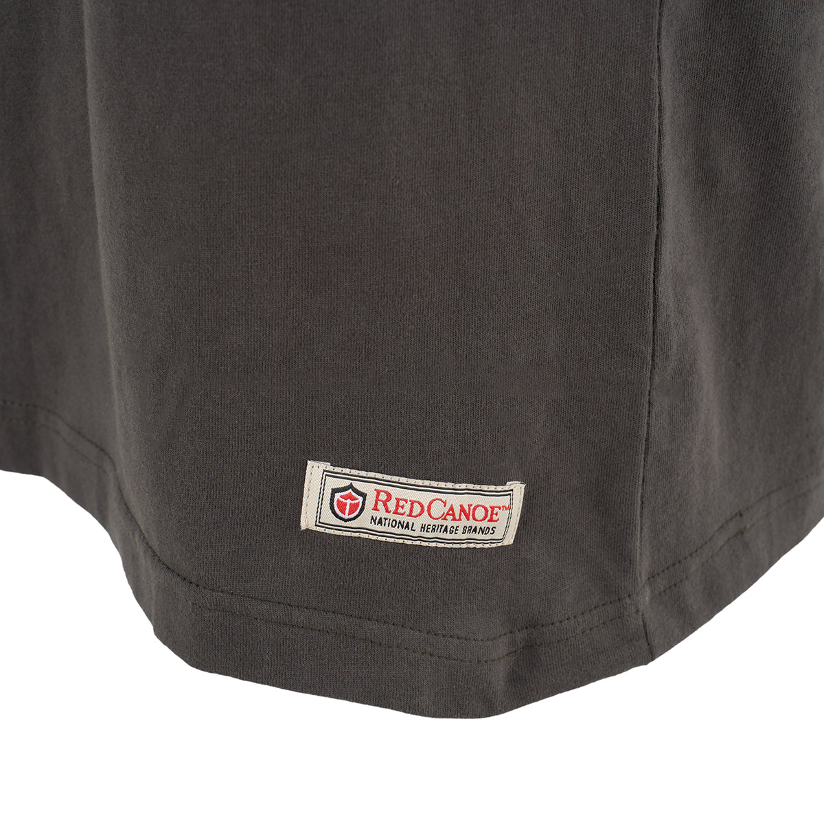 Close-up of the bottom hem of a dark gray Red Canoe Boeing Company T-Shirt from The Boeing Store, showing a rectangular fabric tag with the Red Canoe logo and NATIONAL HERITAGE BRANDS text.