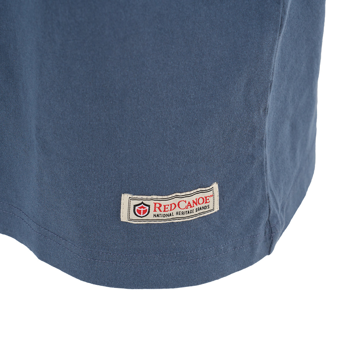 Close-up of the lower hem of a blue Red Canoe NASA Rocket Scientist T-Shirt from The Boeing Store, showing a rectangular patch with a red shield logo and Red Canoe National Heritage Brands text.