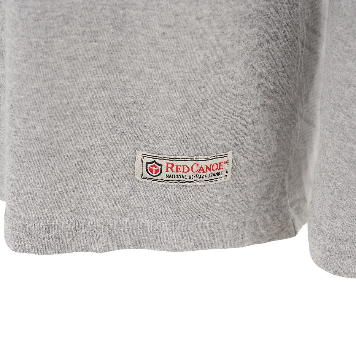 A close-up of the bottom hem of the Red Canoe Boeing Tuskegee Airmen Long Sleeve T-Shirt by The Boeing Store, featuring a rectangular patch inspired by the spirit of the Tuskegee Airmen and P-51 Red Tail Mustang.