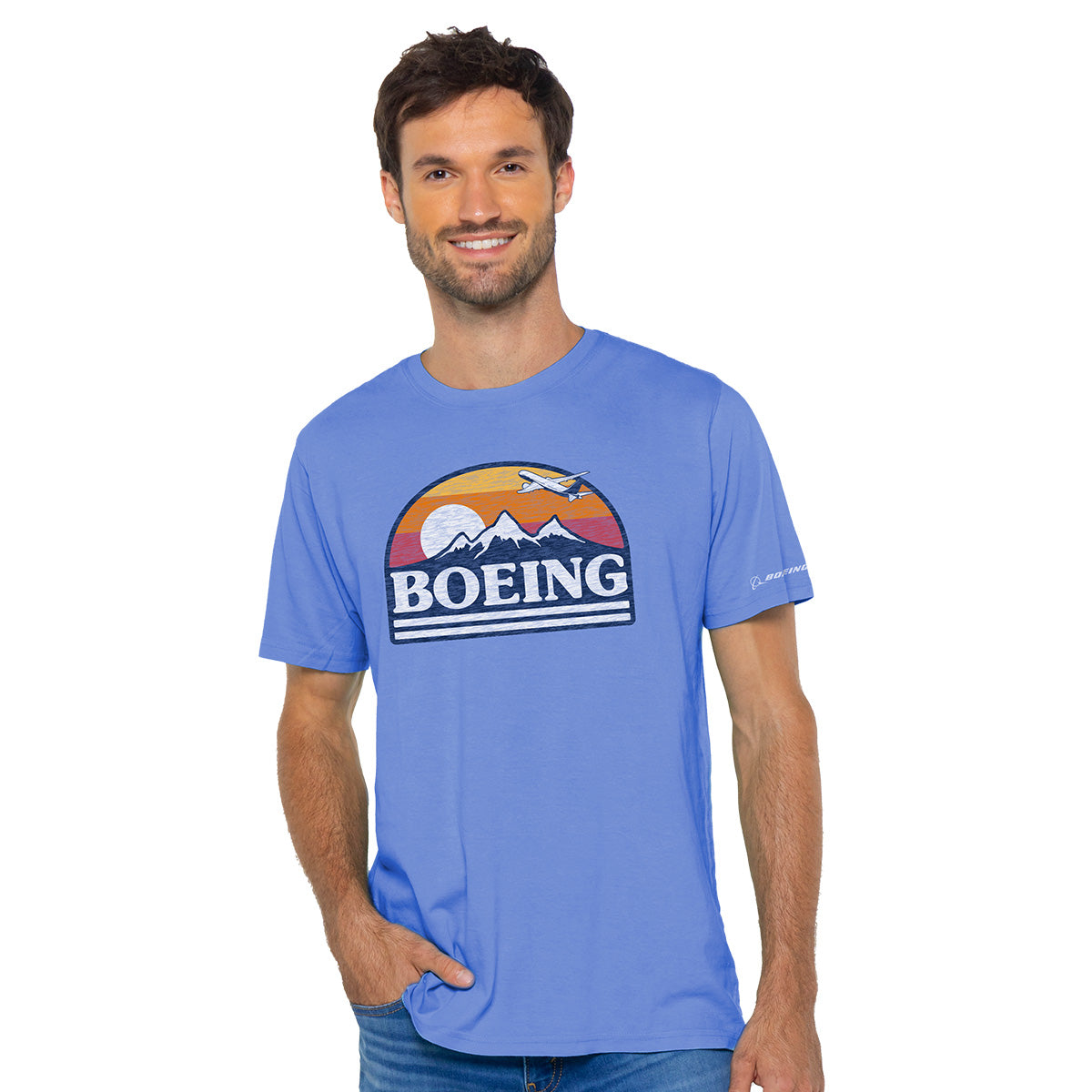 A man with short dark hair and a beard confidently wears a blue Boeing Horizon Mens T-Shirt, showcasing a retro logo with a mountain, sun, and airplane design. With one hand in his pocket against a white background, he displays his passion for aviation.