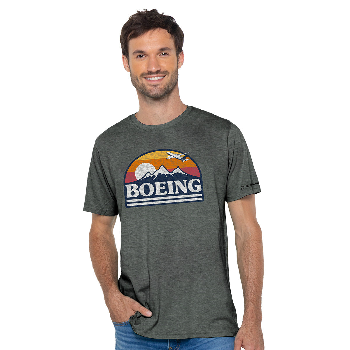 A man confidently smiles with one hand in his jeans pocket, wearing a dark green Boeing Horizon Mens T-Shirt adorned with a design of mountains, a sunset, and an airplane against a plain white backdrop.