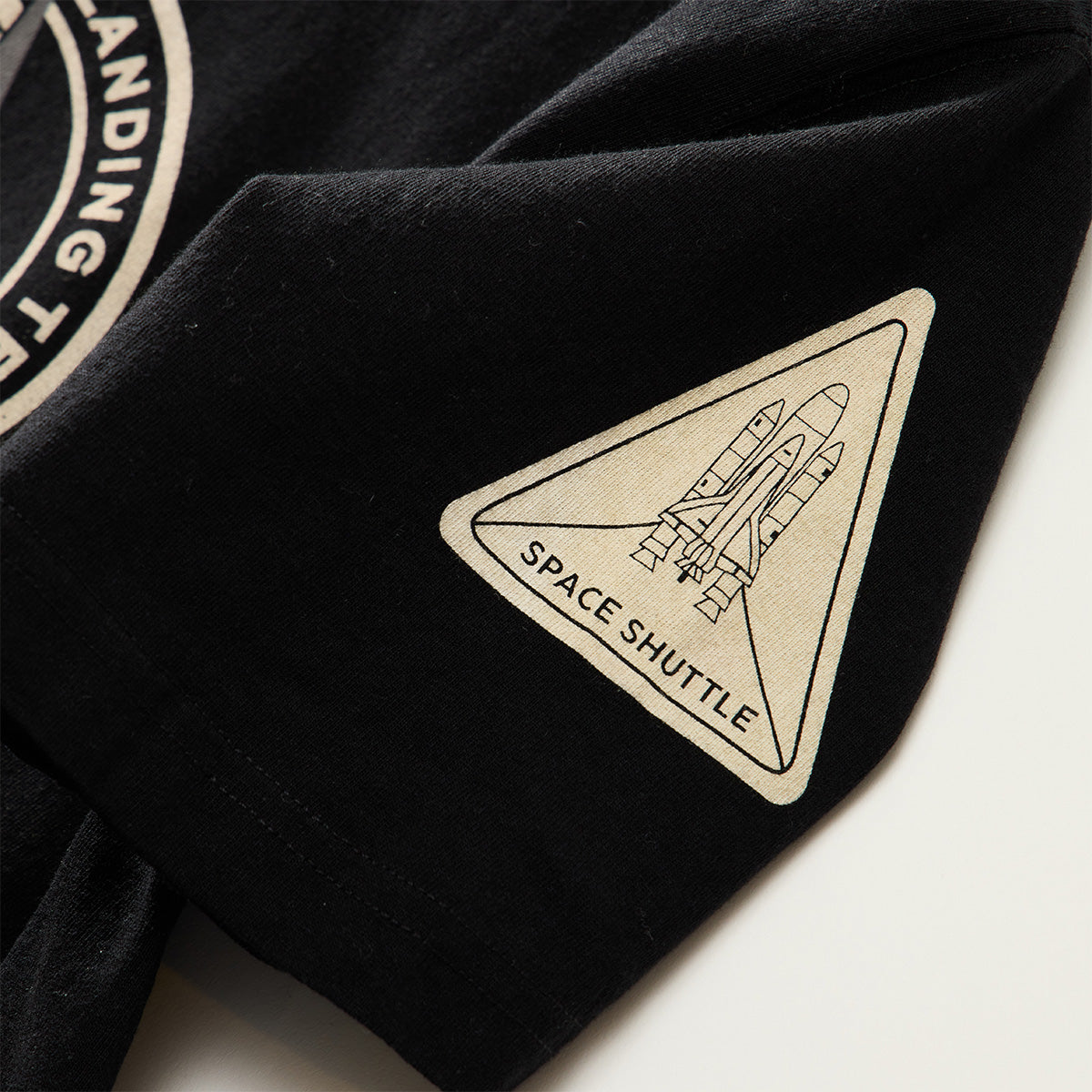 Close-up of a black Red Canoe NASA ALT T-Shirt sleeve by Boeing, featuring a beige triangular patch with a detailed line drawing of a space shuttle and the words SPACE SHUTTLE underneath.