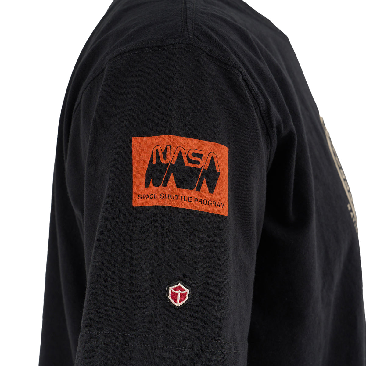 A close-up of the Red Canoe NASA ALT T-Shirt by Boeing shows a black sleeve with an orange Space Shuttle Program patch and a small red and white embroidered emblem below, inspired by the shuttle Approach and Landing Test.