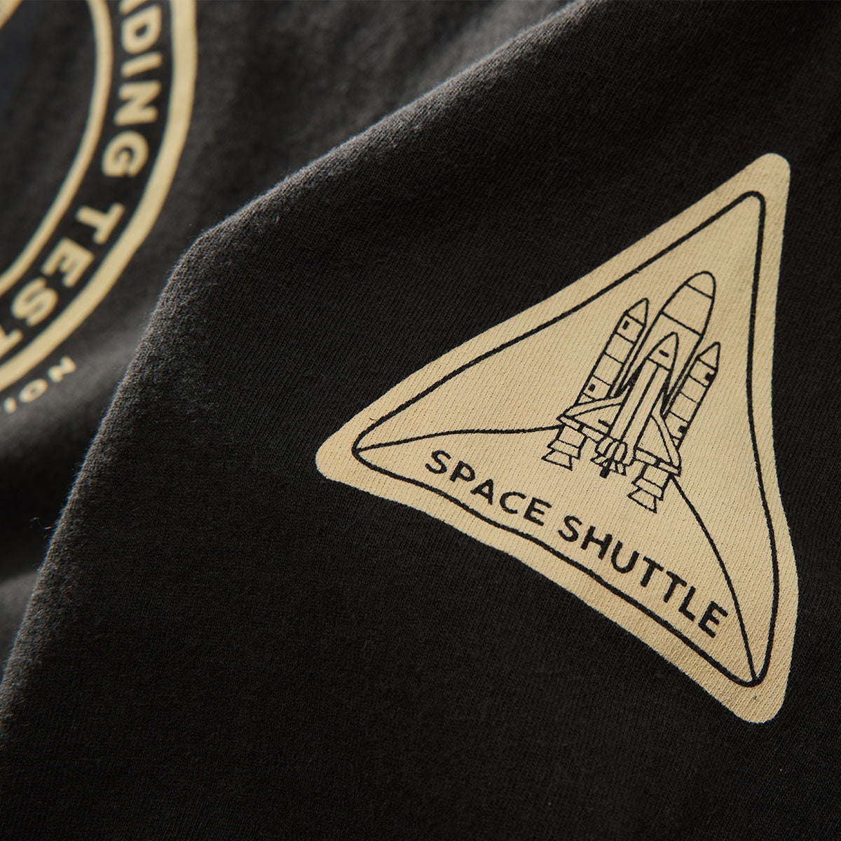 A close-up of a black fabric patch with a beige triangular NASA Space Shuttle emblem and SPACE SHUTTLE text, seen on the BOEING Red Canoe Space Shuttle Mens Long Sleeve T-Shirt. Part of another circular patch is also visible.