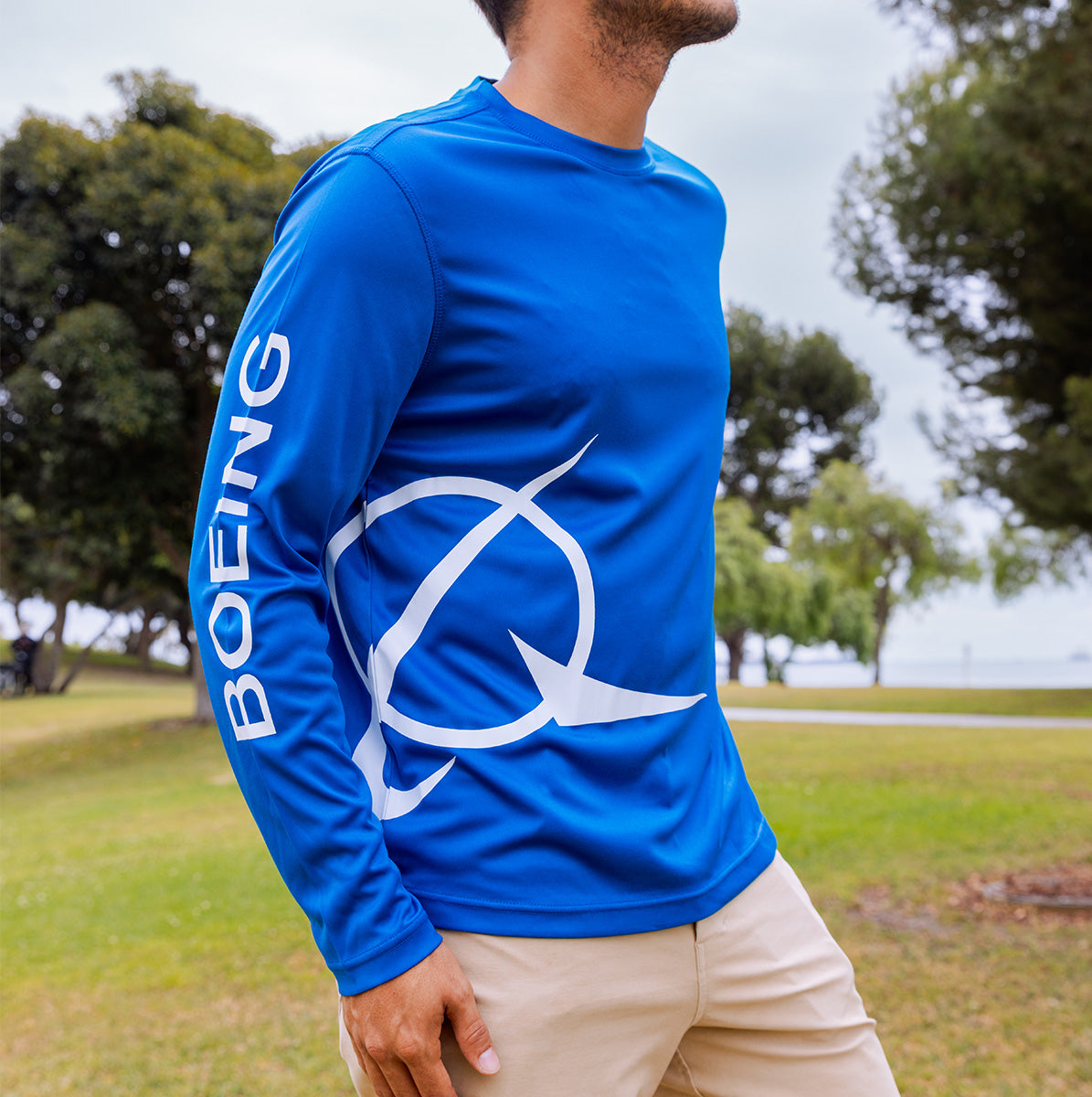 A man wears the Boeing Symbol Logo Men's UV Long Sleeve T-Shirt by BOEING, featuring a bright blue color and logo print down the sleeve, while standing outdoors in a grassy park with trees behind him.