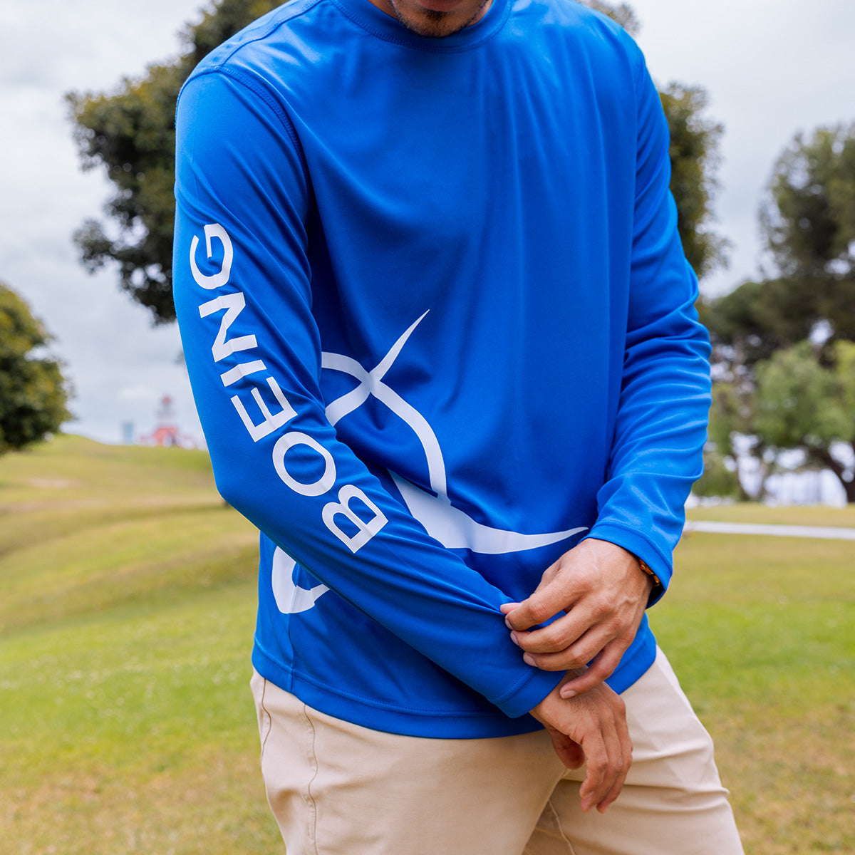 Outdoors, a person adjusts the cuff of a bright blue Boeing Symbol Logo Men's UV Long Sleeve T-Shirt (UPF 50+) by BOEING, featuring "BOEING" and a white logo on the sleeve, with grass and trees in the background.