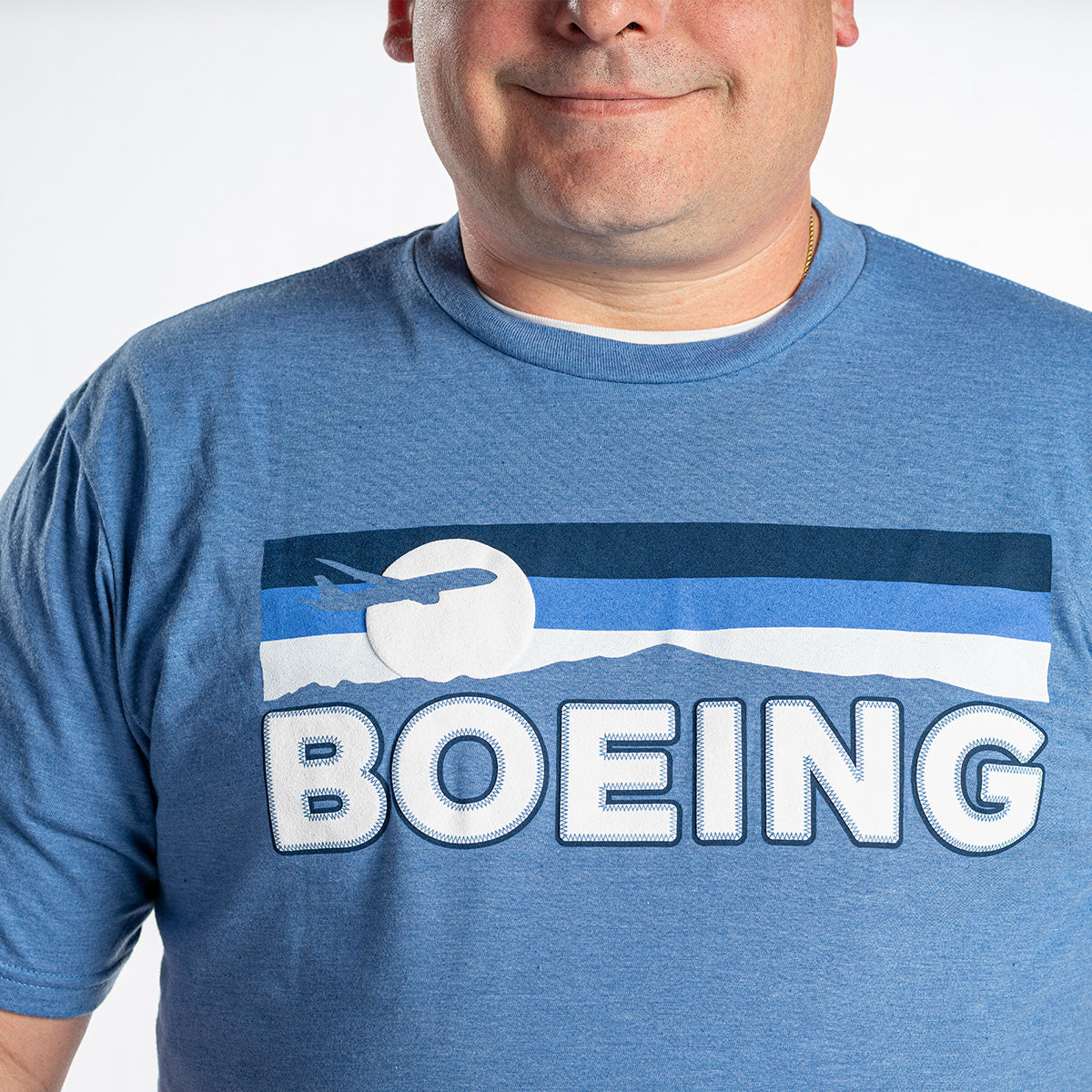 A person wears the Boeing Men's Wanderer T-Shirt by BOEING, showing a retro logo with a stylized airplane and bold white text. Only the lower half of their face and upper torso are visible.