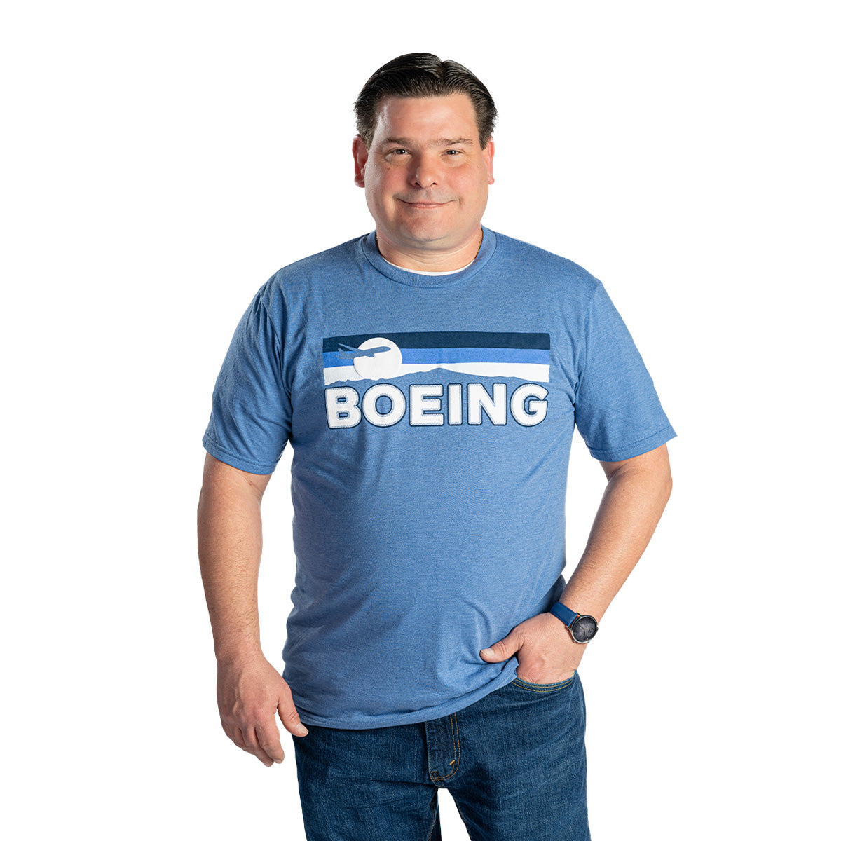 A man wearing a blue Boeing Men's Wanderer T-Shirt by BOEING and jeans stands against a white background, smiling with one hand in his pocket.