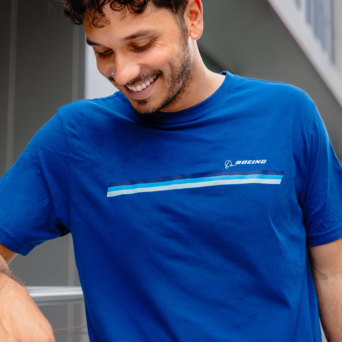 A smiling man with short curly hair wears the Boeing Unisex Signature Logo Stripe T-Shirt by BOEING, a 100% cotton blue tee with horizontal blue and white stripes and the Boeing logo, while standing outdoors near a modern building.