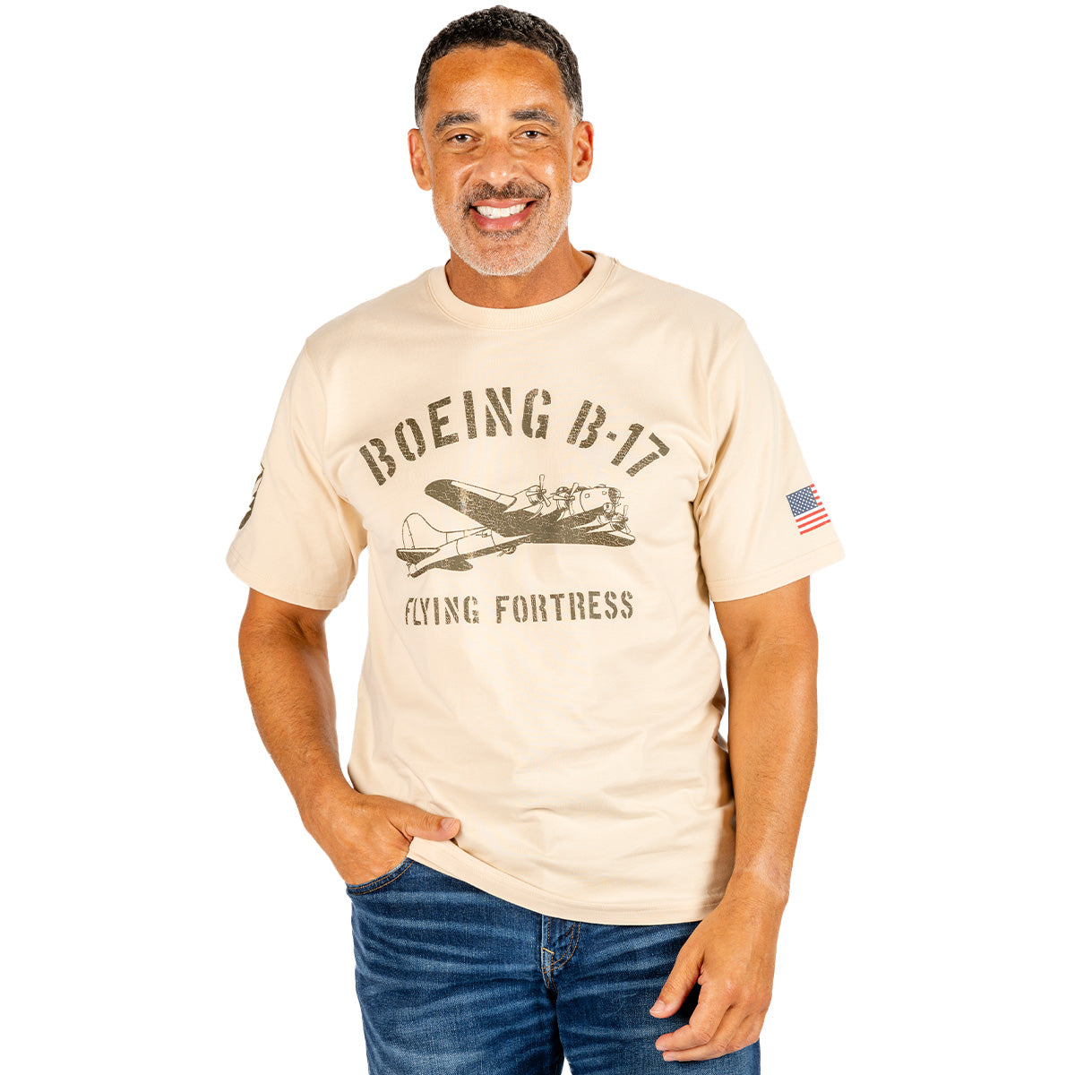 A smiling man models the BOEING Heritage B-17 Flying Fortress Unisex T-Shirt in beige, featuring an authentic airplane graphic and an American flag patch on the sleeve, paired with blue jeans—an ideal style for any aviation enthusiast.
