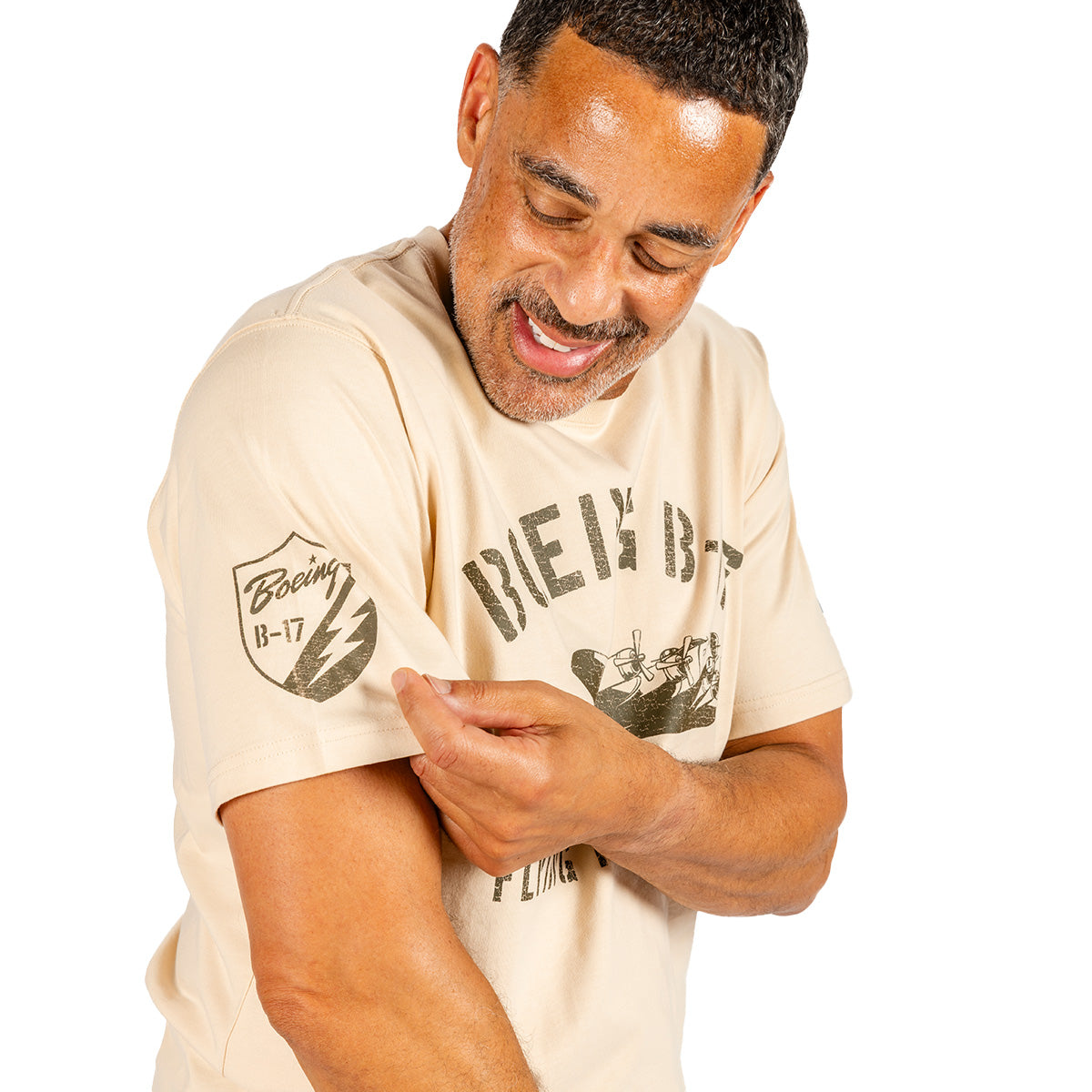 A smiling man in a beige BOEING Heritage B-17 Flying Fortress Unisex T-Shirt points to the distinctive B-17 Flying Fortress patch on his sleeve, proudly displaying his enthusiasm for aviation. The background is plain white.