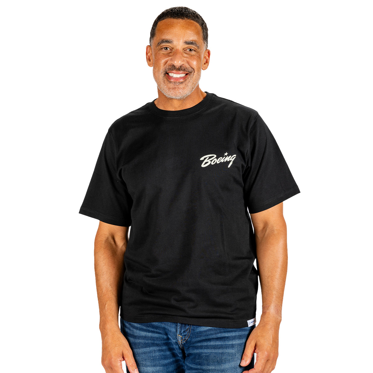 A smiling man stands against a white background, wearing blue jeans and the Boeing Heritage Script B-17 Flying Fortress Unisex T-Shirt by BOEING—a perfect tribute to aviation history.