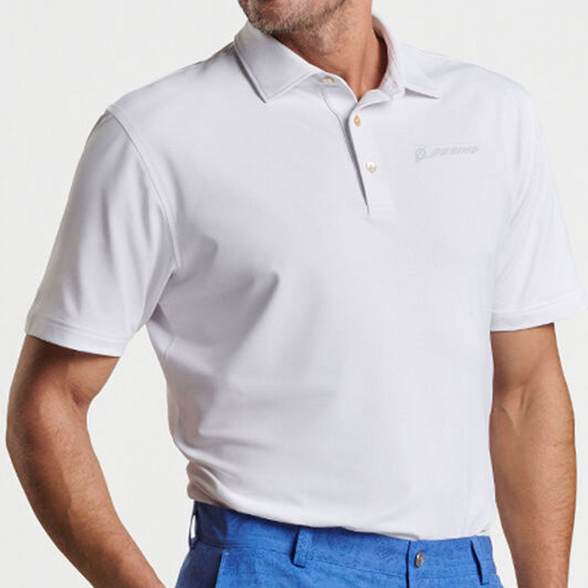 A man wearing the Peter Millar Boeing Men’s Solid Performance Mesh Polo in white, featuring a subtle chest logo, paired with bright blue pants, stands against a plain light background.