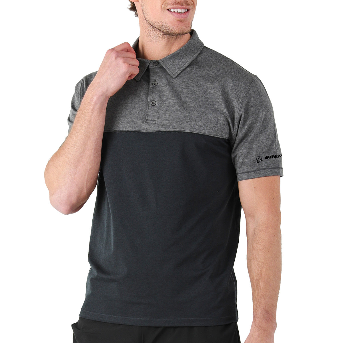 A man with short hair smiles, holding the collar of the Glyder Boeing Signature Halfway Polo by BOEING. The athletic polo features a retro color block design with a dark lower half, lighter gray marbled jersey upper half, and a logo on the left sleeve.