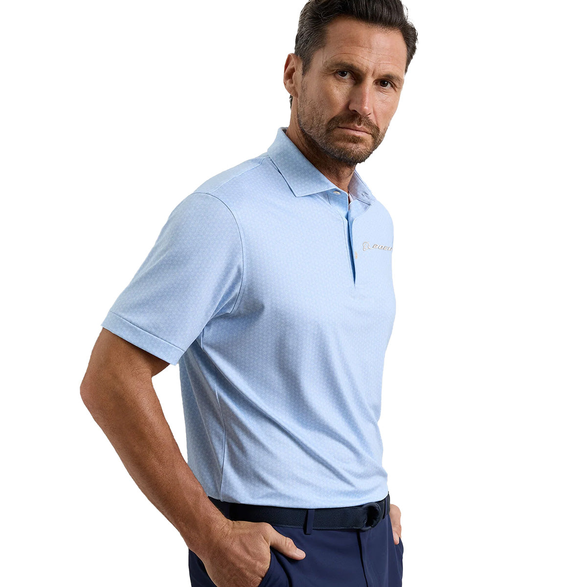 A man with short brown hair and a beard wears a light blue Peter Millar Boeing Men’s Nautilus Performance Polo by BOEING and dark pants, standing with one hand in his pocket against a plain white background.