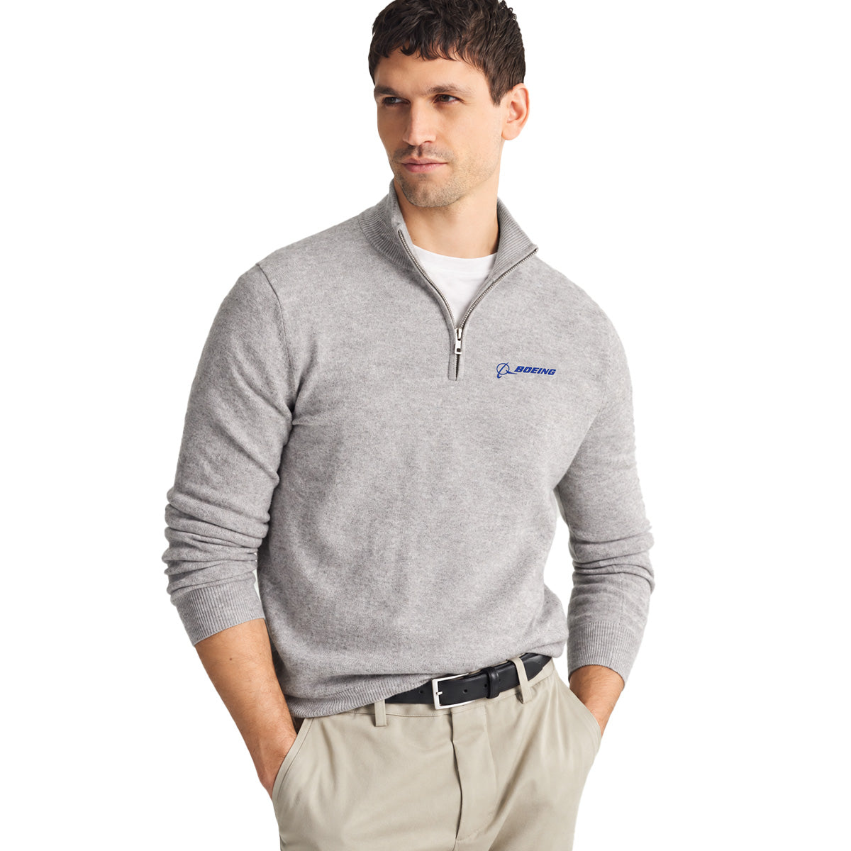 A man wearing the Boeing Signature Men’s Cashmere Quarter-Zip sweater by BOEING in light gray, paired with a white t-shirt, beige pants, and a black belt stands with his hands in his pockets, looking to the side.