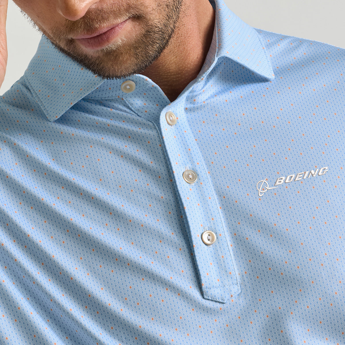 A man wearing a Peter Millar Boeing Men’s Bartlett Performance Polo in light blue, featuring small dot patterns and the Boeing logo on the chest. His face is partially visible as he touches his temple. This BOEING polo offers UPF 50+ sun protection and moisture-wicking technology for all-day comfort.