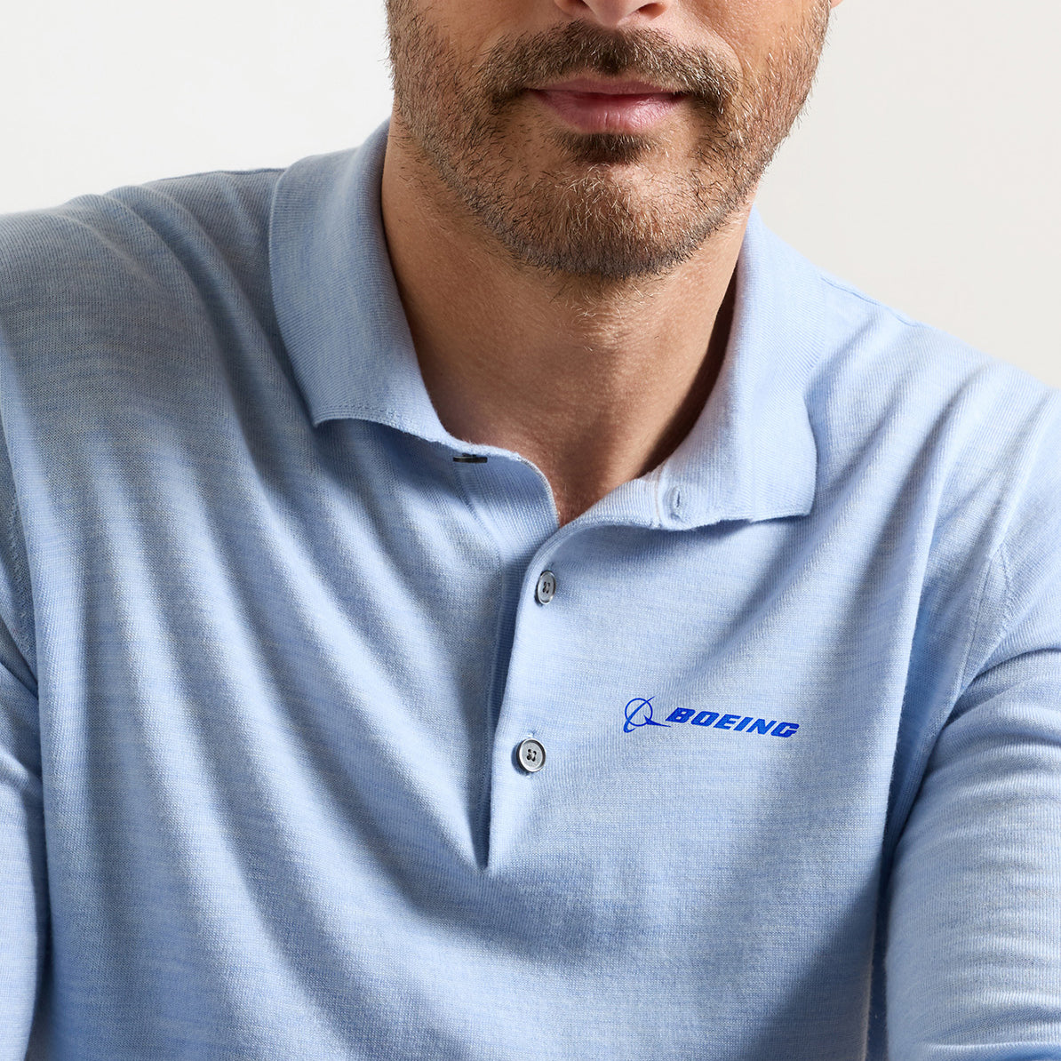 A man is wearing the Peter Millar Boeing Men’s Dorset Sweater Polo, a light blue Merino wool shirt featuring the BOEING logo embroidered on the left chest. His face is partially visible, revealing a neatly trimmed beard and mustache.
