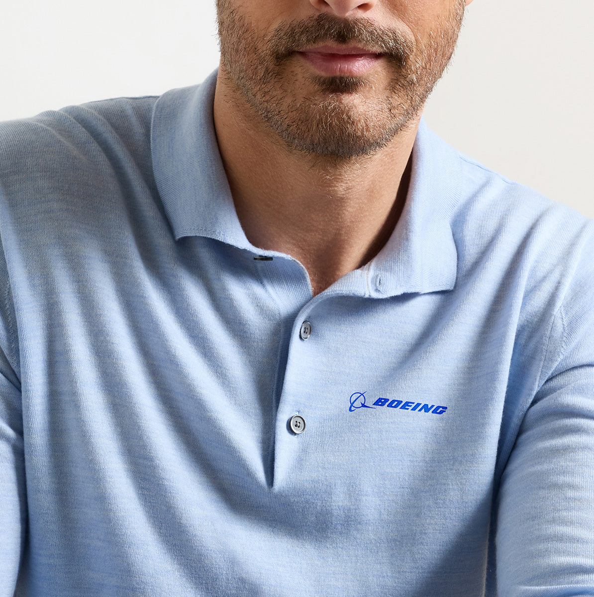 A man is wearing the Peter Millar Boeing Men’s Dorset Sweater Polo, a light blue Merino wool shirt featuring the BOEING logo embroidered on the left chest. His face is partially visible, revealing a neatly trimmed beard and mustache.